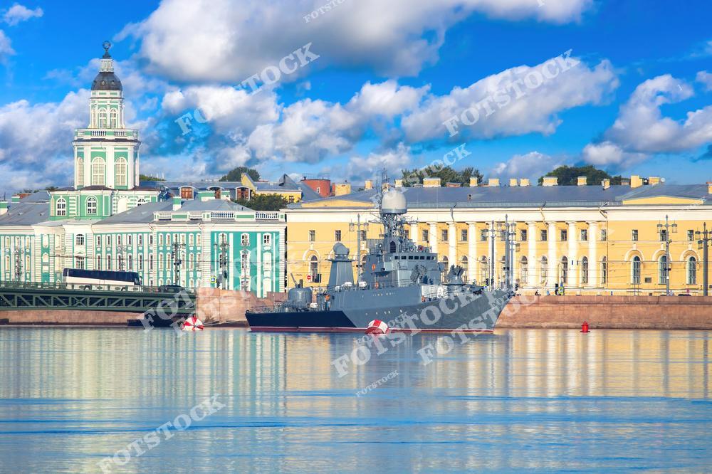 На фотографии изображено военно-морское судно в акватории реки Невы, в центре Санкт-Петербурга. Корабль, находящийся в движении или на якоре, контрастирует с исторической архитектурой города, включая классические здания вдоль набережных. Синева воды и яркое небо создают динамичную атмосферу, подчеркивая важность этого элемента морской мощи России.